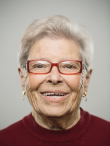 Close up portrait of senior caucasian woman with happy expression against gray white background. Vertical shot of spanish real people smiling in studio with white hair and glasses. Photography from a DSLR camera. Sharp focus on eyes.